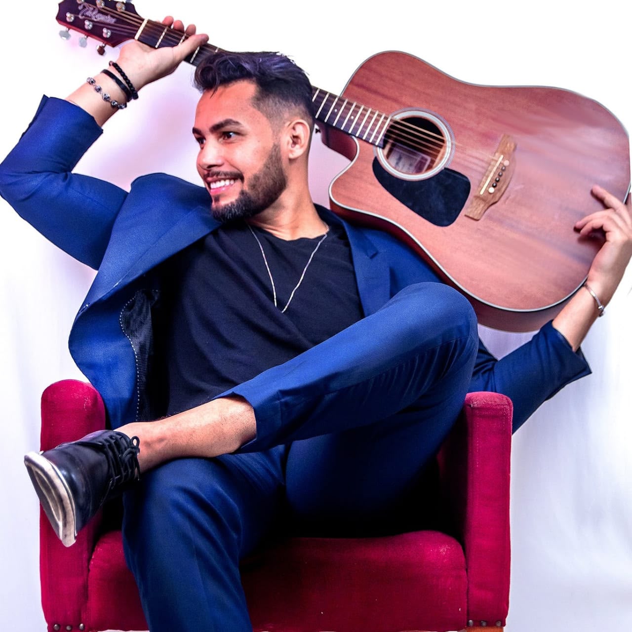 Ricky Moreira sentado em uma cadeira vermelha, usando terno azul e com violão